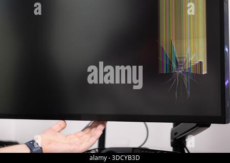 Vue détaillée d'une main humaine présentant un écran de moniteur gravement cassé, présentant des fissures de verre en étoile et des lignes verticales colorées vives d'imp Banque D'Images