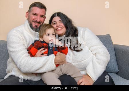 Heureux parents tenant leur tout-petit tout en étant assis ensemble sur un canapé dans un cadre intérieur confortable. Banque D'Images