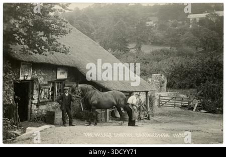 Original début des années 1900 carte postale de charmante scène rurale - un fermier ou un fermier tient son cheval lourd / de travail comme il est attaché par le forgeron / Farrier - à l'extérieur de Branscombe Village Smithy / Forge, Devon, Angleterre, forgeron victorien Royaume-Uni. Cette forge du XVIIIe siècle est la plus ancienne forge de travail au toit de chaume en Angleterre. Vers 1920 Banque D'Images