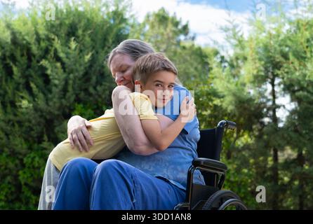 Grand-mère en fauteuil roulant embrassant petit-fils, exprimant l'amour et le lien familial au parc Banque D'Images