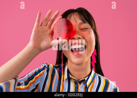 Beau portrait de femme chinoise sur fond coloré. Femme asiatique joyeuse avec une chemise rayée vibrante et des boucles d'oreilles, riant en regardant à travers Banque D'Images