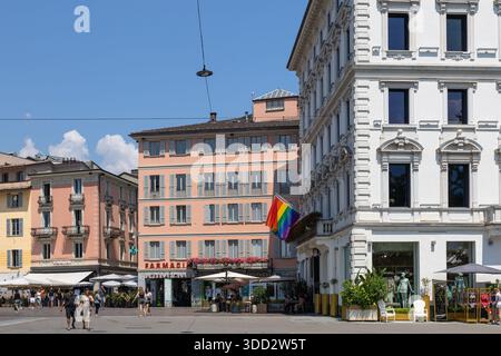 LUGANO, SUISSE, 06.20.2025:place ensoleillée de la ville européenne avec un bâtiment rose pâle, une grande façade blanche, des cafés en plein air, des piétons et un promi Banque D'Images