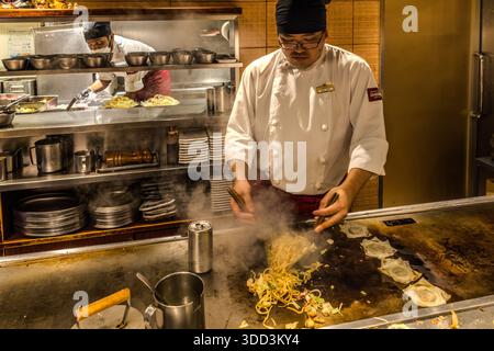 Un chef fait frire des nouilles sur la plaque de cuisson Teppan d'un restaurant à Osaka, au Japon, tandis qu'un deuxième chef prépare des portions d'okonomiyaki en arrière-plan. La vapeur s'élève des nouilles, et une canette de boisson se trouve au bord de la plaque. Le plat est du yakisoba – nouilles de blé frites avec du chou et des légumes – et figure parmi les plats de cuisson les plus populaires d'Osaka, aux côtés de l'okonomiyaki. Banque D'Images