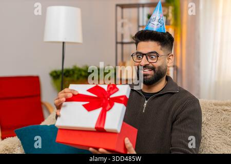 Homme indien à la maison ouvre heureux cadeau d'anniversaire tout en étant assis sur le canapé avec le sourire et l'excitation. Arabian Guy semble surpris et joyeux, ravi par un Banque D'Images