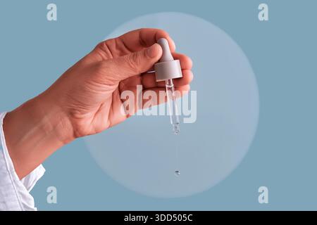 Main tenant une pipette en verre, distribuant une goutte liquide claire de sérum cosmétique. Concentrez-vous sur la routine beauté et les soins naturels de la peau Banque D'Images