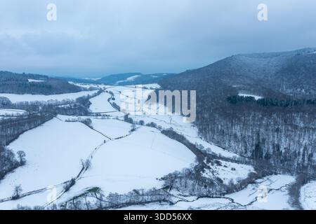 Vue aérienne d'une vallée enneigée sinueuse bordée d'une forêt d'hiver dense et de collines ondulantes dans la région du Morvan. Ciel couvert et tons bleus doux évoquent un paysage hivernal paisible et isolé. Banque D'Images