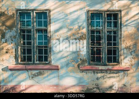 Deux vieilles fenêtres en bois sur le mur de pelage avec des fissures et des ombres comme fond de décomposition urbaine Banque D'Images