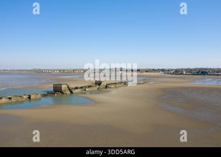 Vue aérienne des vestiges fragmentés du port en béton partiellement submergés dans des bassins peu profonds sur de vastes étendues sablonneuses à marée basse près d'Asnelles, en Normandie, avec un village lointain et un ciel bleu clair. Banque D'Images