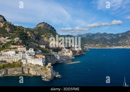 Une vue aérienne panoramique capture les bâtiments blanchis à la chaux d'Amalfi, en Italie, descendant en cascade des falaises escarpées vers la mer Méditerranée d'un bleu vif. Le littoral spectaculaire est encadré par des collines en terrasses, une végétation luxuriante et des montagnes lointaines sous un ciel clair. Banque D'Images