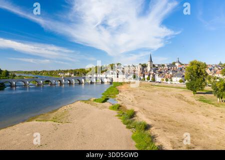 Vue imprenable sur la Charite sur Loire avec son pont de pierre historique traversant la Loire, ses berges sablonneuses au premier plan et la flèche de l'église de la ville s'élevant au-dessus des toits traditionnels. Des nuages tortueux s'étendent sur un ciel bleu vif, soulignant le cadre tranquille au bord de la rivière dans le centre de la France. Banque D'Images