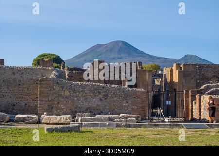 Les murs de pierre altérés et les restes d'une maison romaine se dressent sous l'imposante silhouette du Vésuve à Pompéi, en Italie. La scène est baignée de lumière du jour, mettant en évidence les textures des vestiges archéologiques contre le paysage volcanique. Banque D'Images