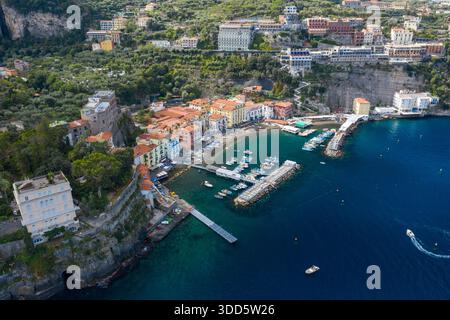 Panorama à grand angle de Sorrente, en Italie, avec des maisons vibrantes regroupées autour d'un petit port de plaisance et des falaises spectaculaires s'élevant derrière. La scène présente une eau méditerranéenne turquoise, une végétation luxuriante et une atmosphère côtière animée. Banque D'Images