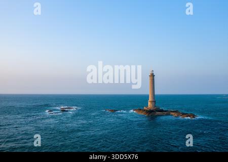 Un grand phare se dresse seul sur un petit éperon rocheux entouré par une mer bleu profond au Cap de la Hague. La scène est éclairée par la lumière du jour claire, avec des vagues douces et un vaste ciel ouvert créant un sentiment d'isolement et de force maritime. Banque D'Images