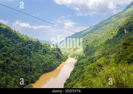 Une rivière boueuse sinueuse coule à travers une vallée abrupte et densément boisée sous un ciel bleu vif dans le nord du Vietnam. Des pentes verdoyantes et des nuages épars créent un paysage naturel et vibrant. Banque D'Images