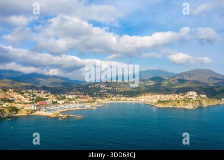 Panorama aérien de Banyuls-sur-mer montrant une ville côtière animée, un port de plaisance abrité et des montagnes spectaculaires sous un ciel parsemé de nuages et de lumière douce du soleil. Banque D'Images