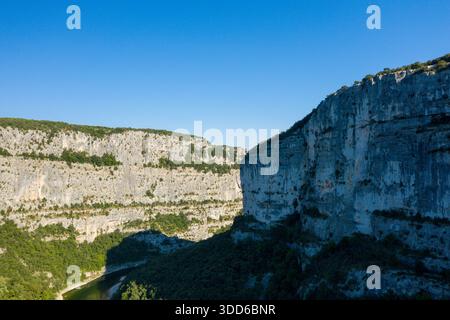 Des murs de calcaire massifs s'élèvent abruptement au-dessus d'une rivière sinueuse dans les Gorges de l'Ardèche, avec une forêt verte dense couvrant le fond de la vallée sous un ciel bleu clair. Le contraste spectaculaire de la lumière et de l'ombre met en évidence les formations rocheuses accidentées. Banque D'Images