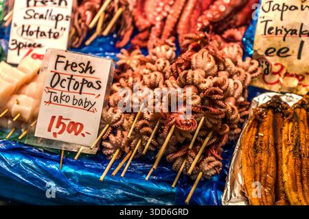Poulpe bébé frais embroché pour la vente au marché Kuromon Ichiba à Osaka, Japon (officiellement licencié depuis 1902, ses racines en tant que lieu de commerce remontant au début du XIXe siècle). Un panneau fait la promotion de « Fresh Octopas Tako Baby » pour 1 500 yens en anglais, avec des pétoncles et de l’anguille en sauce teriyaki exposés à côté. Banque D'Images
