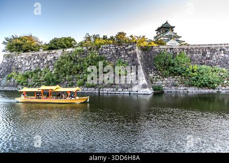 Un bateau de tourisme de style traditionnel transporte des touristes le long du fossé d’Osaka Castle à Osaka, au Japon, sa tour principale visible au-dessus du mur de pierre. Toyotomi Hideyoshi a fait construire le château en 1583. Après plusieurs destructions, la tour principale actuelle a été reconstruite en béton armé en 1931. Banque D'Images