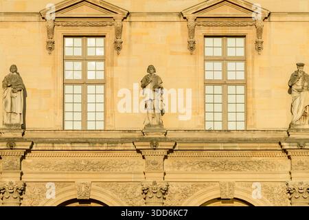 Trois statues de pierre classiques se dressent entre de hautes fenêtres sur la façade ornée du palais du Louvre à Paris, en France. La lumière dorée met en valeur les reliefs complexes, l'architecture symétrique et les détails raffinés de la maçonnerie. Banque D'Images