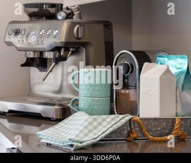 Une station de café maison moderne disposée sur un comptoir de cuisine, avec une machine à expresso en acier inoxydable, un moulin à café, des tasses en céramique et des accessoires soignés. L'image transmet une routine matinale calme et une vie domestique contemporaine, avec des tons neutres et des éléments de design épurés. Idéal pour les concepts liés au style de vie, aux intérieurs, au marketing immobilier, à la culture du café et au confort quotidien. Photographié avec une faible profondeur de champ pour mettre en valeur la texture et les détails tout en conservant une atmosphère chaleureuse et accueillante. Banque D'Images