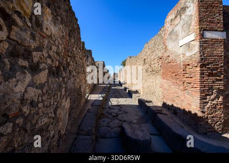 Longue vue sur une étroite rue romaine bordée de murs en pierre altérée et en briques dans le site archéologique de Pompéi. L'image présente une maçonnerie brute, des ombres profondes et l'aménagement urbain historique sous un ciel bleu vif. Banque D'Images