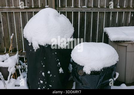 La neige recouvre des poubelles noires dans une cour arrière de banlieue après une tempête hivernale tôt le matin. Banque D'Images