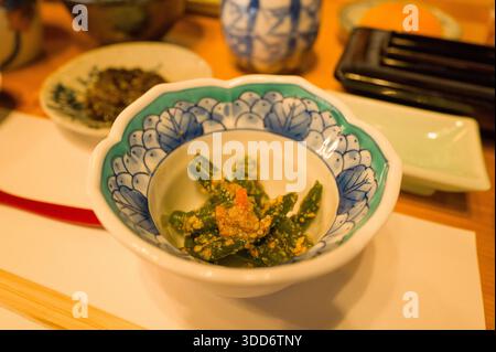 Les haricots verts enrobés de sauce au sésame sont servis dans un bol en porcelaine bleue et blanche orné, qui fait partie d'un petit-déjeuner japonais traditionnel servi sur une table en bois avec un éclairage ambiant chaleureux. Banque D'Images