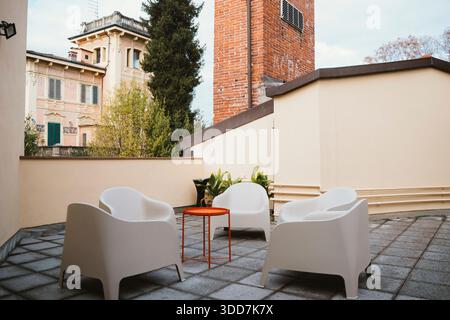 Terrasse extérieure avec chaises blanches modernes et une table orange sur fond d'architecture historique et de verdure, créant un cadre relaxant Banque D'Images