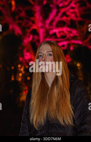 Cette photographie représente une jeune femme aux cheveux roux saisissants, placée sur un fond éclatant de lumières de Noël roses et rouges. La femme porte un Banque D'Images