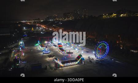 Parc d'attractions lumineux avec une grande roue et des manèges colorés illuminant la nuit au bord de la mer Banque D'Images