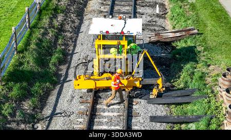 Les ouvriers de la construction sont occupés à réparer les voies ferrées. Une machine lourde les assiste dans la tâche. La lumière du soleil brille sur la scène avec de l'herbe verte à proximité. Banque D'Images