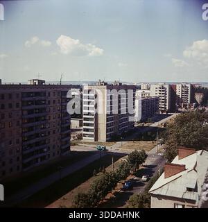 Slavyansk, Ukraine, URSS, années 1980 Une vue panoramique à angle élevé du quartier résidentiel nouvellement construit sur la rue Yunych Komunariv (maintenant rue Bankivska). Plusieurs blocs d'appartements massifs de 9 étages en briques grises avec des rangées de balcons dominent le paysage. Une camionnette soviétique bleue longe la large avenue pavée bordée de jeunes arbres en herbe. Au premier plan, le toit en ardoise d'un bâtiment plus ancien contraste avec les gratte-ciel modernes. Au loin, des montagnes de craie industrielle blanche sont visibles à l'horizon. Banque D'Images