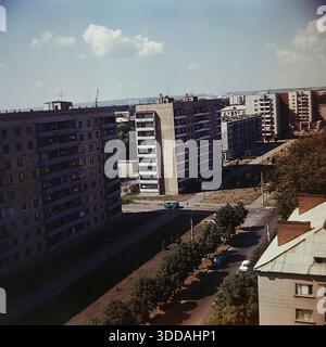 Slavyansk, Ukraine, URSS, années 1980 Une vue panoramique à angle élevé du quartier résidentiel nouvellement construit sur la rue Yunych Komunariv (maintenant rue Bankivska). Plusieurs blocs d'appartements massifs de 9 étages en briques grises avec des rangées de balcons dominent le paysage. Une camionnette soviétique bleue longe la large avenue pavée bordée de jeunes arbres en herbe. Au premier plan, le toit en ardoise d'un bâtiment plus ancien contraste avec les gratte-ciel modernes. Au loin, des montagnes de craie industrielle blanche sont visibles à l'horizon. Banque D'Images