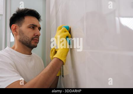 Homme barbu concentré nettoyant les carreaux de salle de bain avec une éponge bleue tout en portant des gants en caoutchouc jaune, en se concentrant sur le nettoyage du mur carrelé blanc pour Banque D'Images