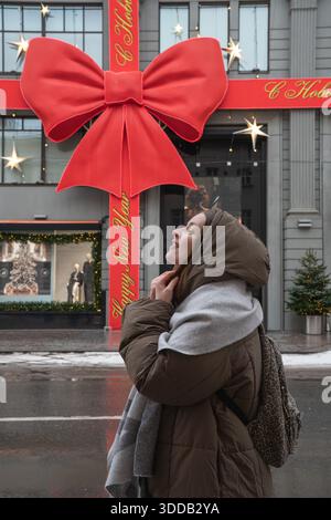 Moscou, Russie - 28 décembre 2025 : femme debout sur la rue humide devant la façade du grand magasin TSUM décorée d'un grand arc rouge, nouvel an Banque D'Images