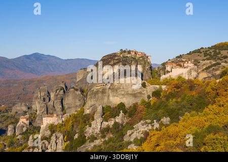 Vue panoramique sur les Météores en Grèce, avec d'anciens monastères perchés sur d'imposantes falaises de grès entourées de feuillage d'automne et de montagnes lointaines sous un ciel bleu clair. Le paysage met en valeur des formes géologiques saisissantes et des couleurs saisonnières vibrantes. Banque D'Images