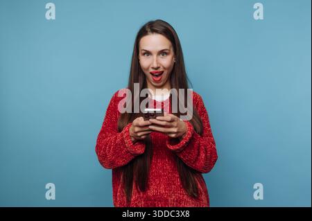 Femme en pull rouge regardant surpris tout en utilisant un smartphone. Banque D'Images