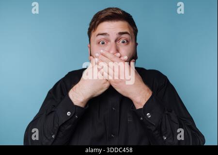 Un homme surpris couvre sa bouche avec les deux mains sur un fond bleu. Banque D'Images