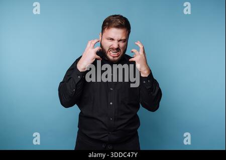 Homme en chemise noire exprimant la colère sur fond bleu. Banque D'Images