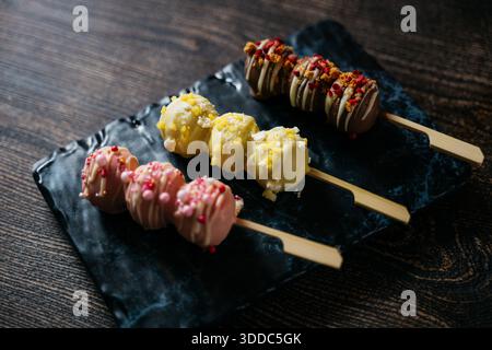Trois desserts colorés différents sont affichés sur une plaque noire. Les desserts sont décorés de saupoudrées et sont servis sur des bâtonnets de bambou. La plaque Banque D'Images