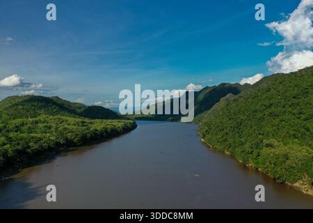 Un large tronçon du fleuve Mékong coule entre des collines verdoyantes densément boisées sous un ciel bleu vif avec des nuages éparpillés. La scène présente une végétation tropicale riche, des pentes douces et une eau tranquille en Asie du Sud-est. Banque D'Images