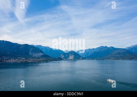 Un ferry blanc glisse à travers l'étendue calme du lac majeur dans le nord de l'Italie, avec en toile de fond des montagnes escarpées et des nuages sinueux qui s'étendent sur un ciel lumineux. La scène capture le vaste lac, le rivage lointain et le paysage alpin tranquille. Banque D'Images