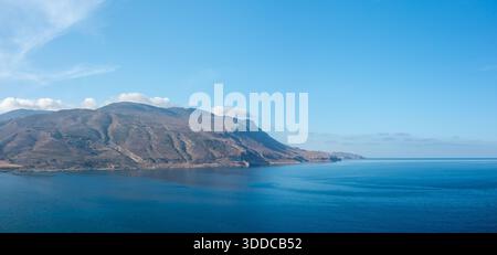 Une étendue tranquille de mer bleu profond rencontre des montagnes escarpées et ensoleillées à pointe de Balos, en Crète. Le ciel ouvert et l'eau douce créent une scène méditerranéenne paisible et expansive. Banque D'Images