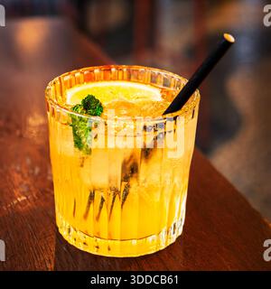 Un cocktail ambré rafraîchissant avec une tranche d'orange et de menthe, servi dans un verre avec une paille noire sur une surface en bois. Banque D'Images