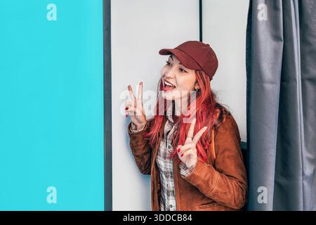 Jeune femme souriante, faisant des signes de paix et montrant une expression vibrante à l'intérieur d'une cabine photo pour des souvenirs amusants Banque D'Images