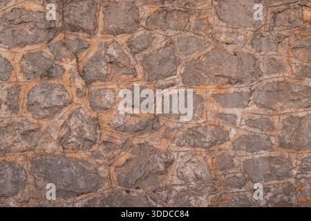 Un mur de pierre accidenté composé de rochers irréguliers et gris liés par un mortier épais de couleur beige. La surface altérée présente des fissures naturelles et minérales Banque D'Images