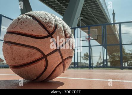 Bangkok, Thaïlande - 18 décembre 2025 - Un ballon de basket-ball très utilisé repose sur un terrain extérieur, avec un cerceau et un panneau en fibre de verre visibles sous l'indus Banque D'Images