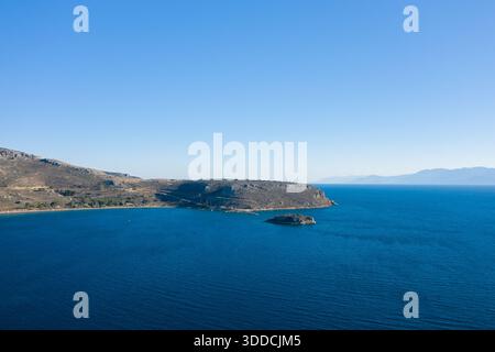 Une vue panoramique sur une péninsule escarpée et une petite île entourée d'une eau d'un bleu profond, avec des montagnes lointaines et un ciel sans nuages créant un sentiment d'ouverture et de calme. La côte ensoleillée présente des textures terreuses et une lumière méditerranéenne tranquille. Banque D'Images