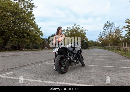 Jeune femme debout près de sa moto dans un parking, tenant un casque orange Banque D'Images