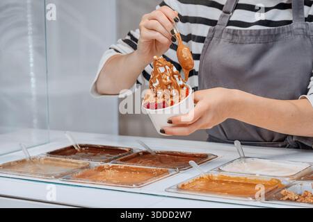 Employé préparant une tasse de yogourt glacé frais avec des fraises et de la sauce au chocolat fondu dans un magasin de desserts Banque D'Images
